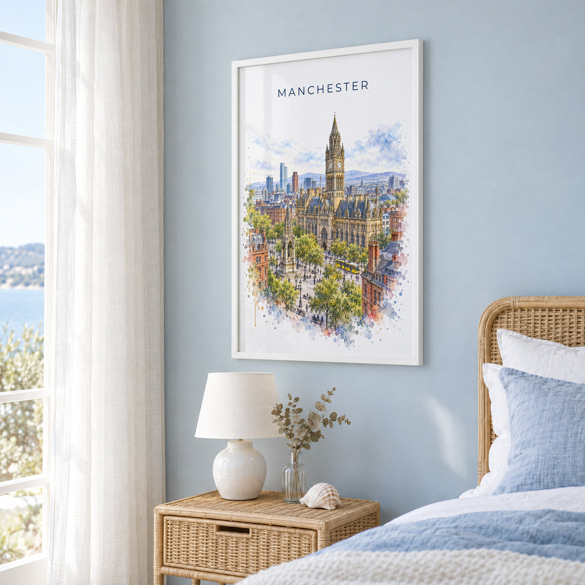 Manchester, England watercolor print in a white frame on a pale blue wall beside a rattan bed in a coastal bedroom