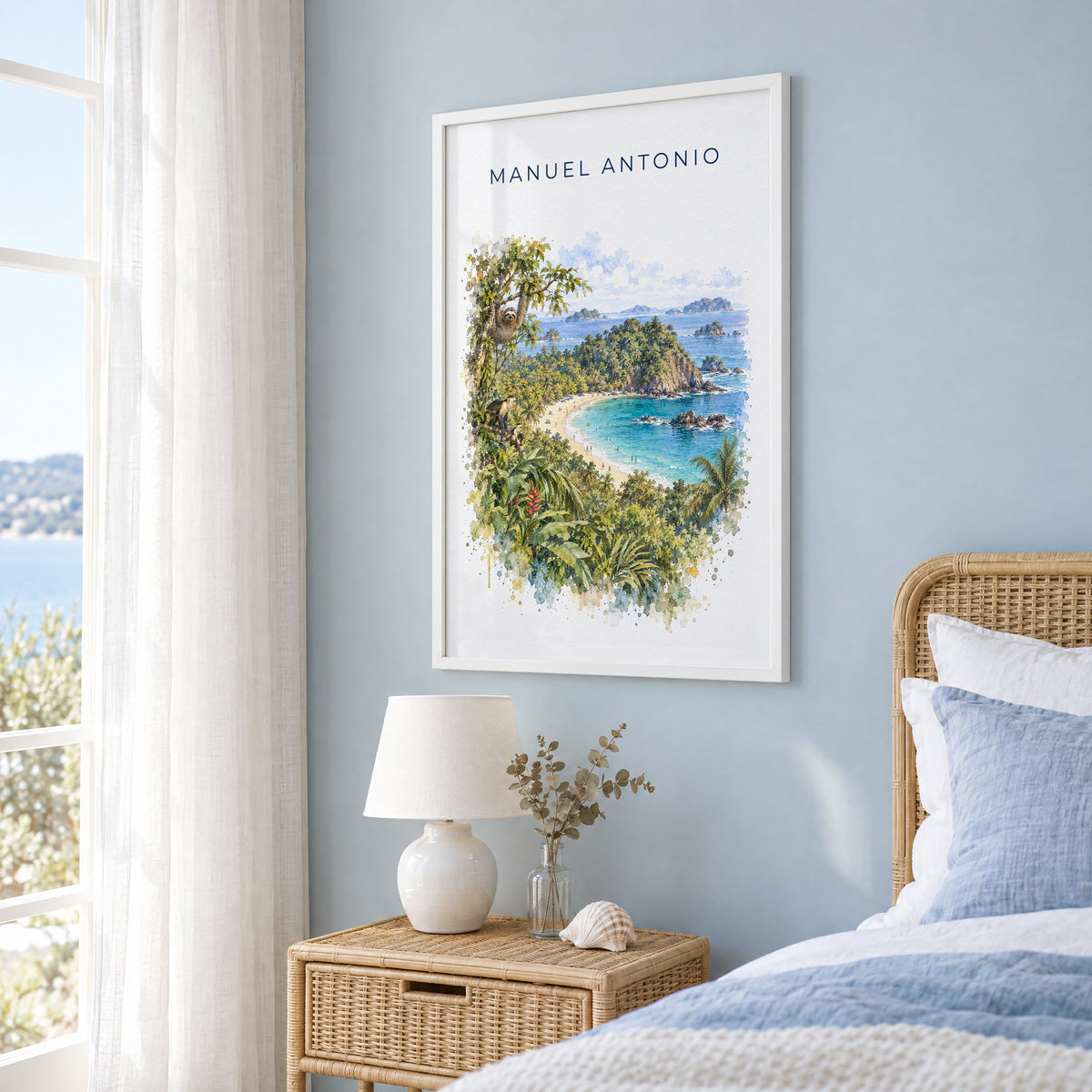Manuel Antonio watercolor print in a white frame on a pale blue wall beside a rattan bed in a coastal bedroom