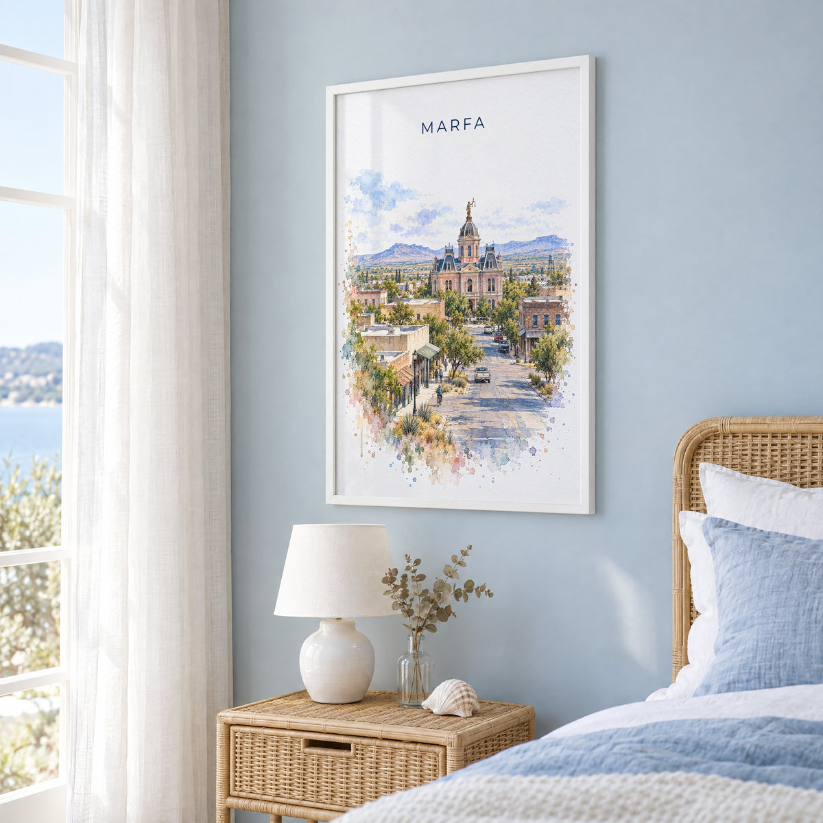 Marfa watercolor print in a white frame on a pale blue wall beside a rattan bed in a coastal bedroom