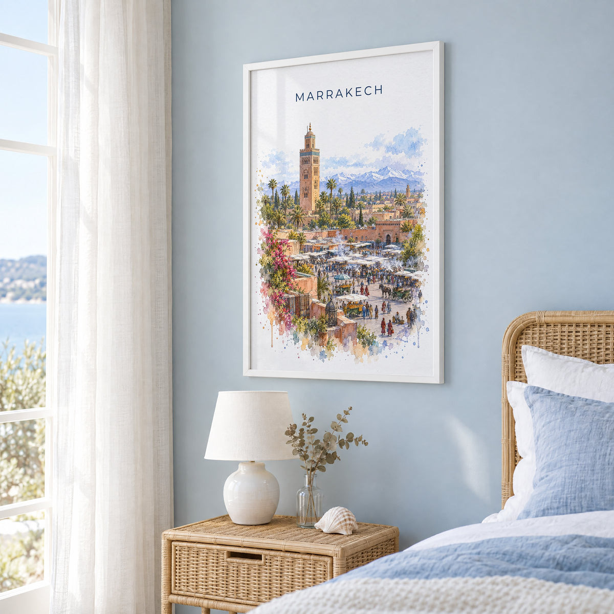 Marrakech watercolor print in a white frame on a pale blue wall beside a rattan bed in a coastal bedroom