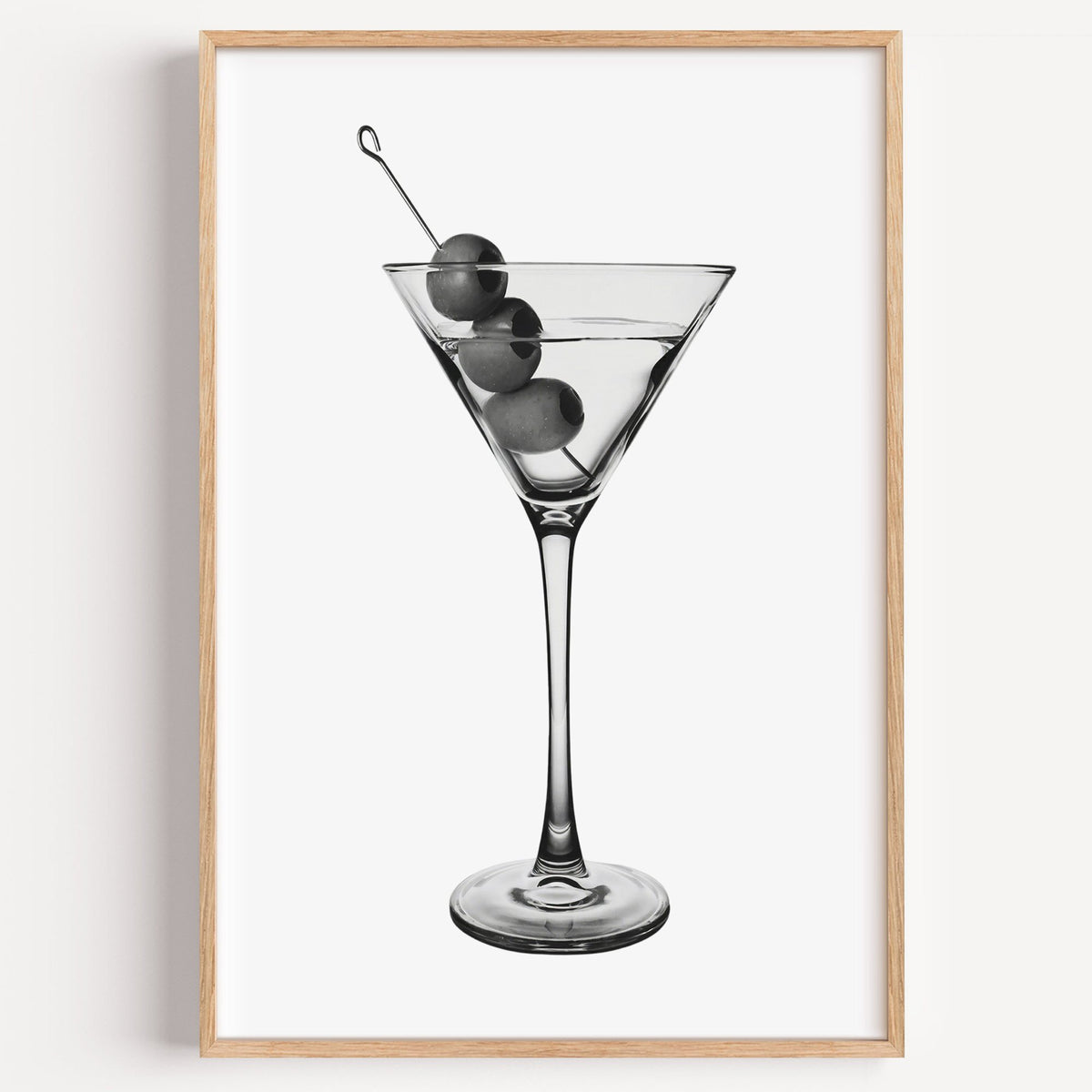 Martini glass with three olives on white print in a light oak frame on a plain white wall