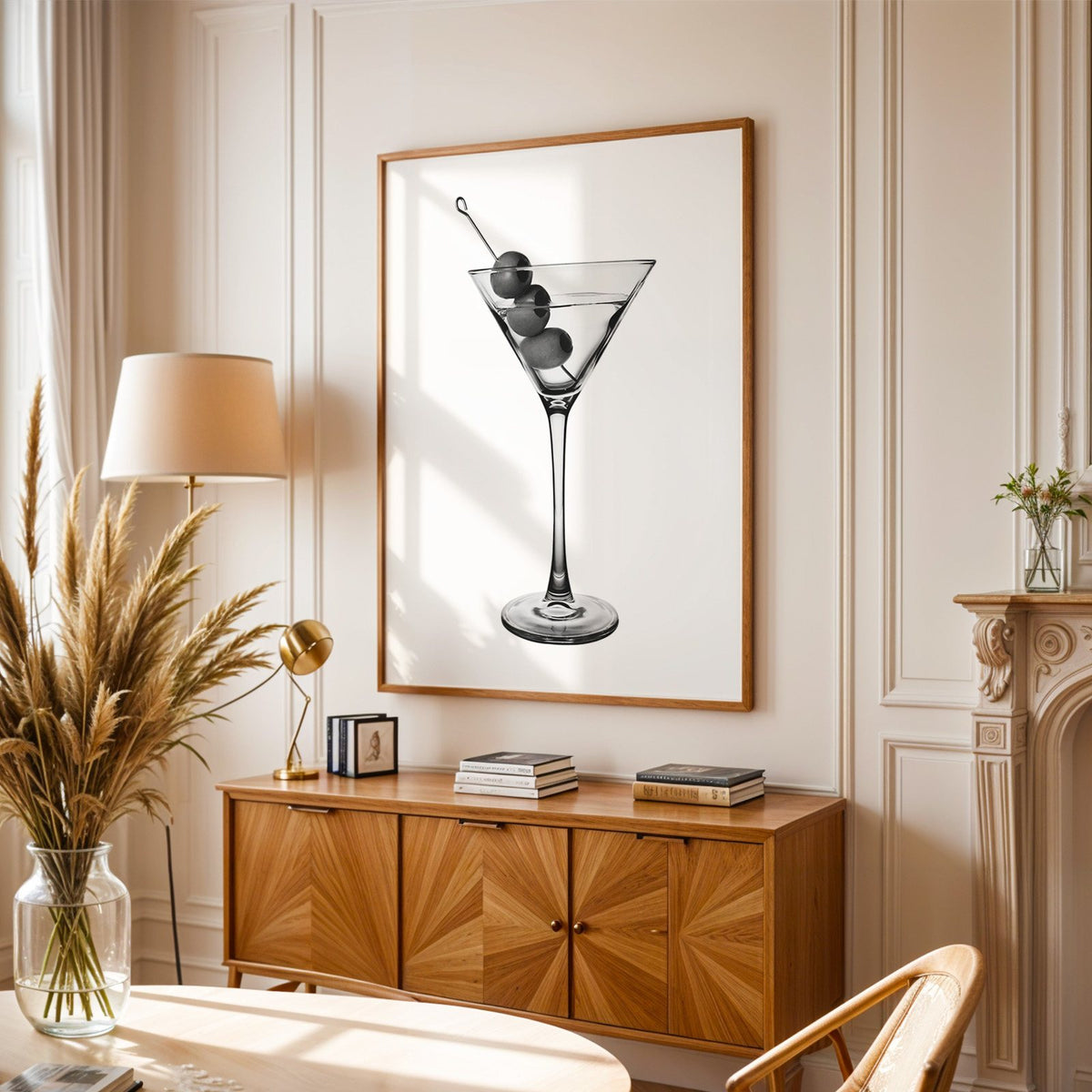 Martini glass with three olives on white print in an oak frame above a sunburst wood sideboard with pampas grass