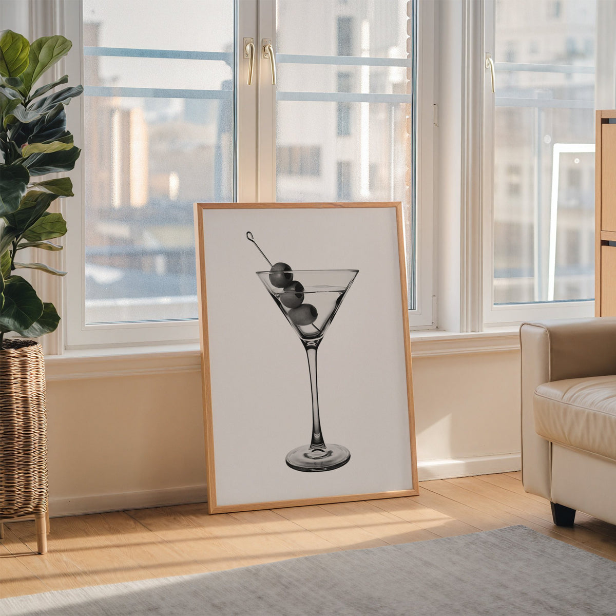 Martini glass with three olives on white print in an oak frame leaning on the floor beside a fiddle leaf fig