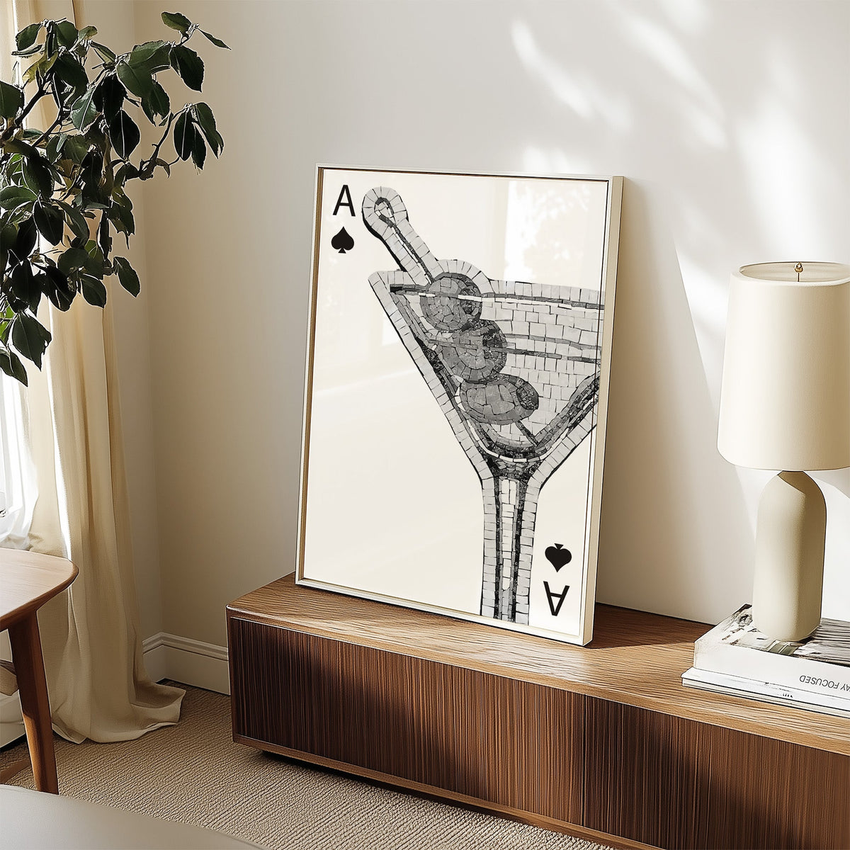 Mosaic martini glass on an ace of spades card print in a white frame on a walnut sideboard beside a ceramic lamp