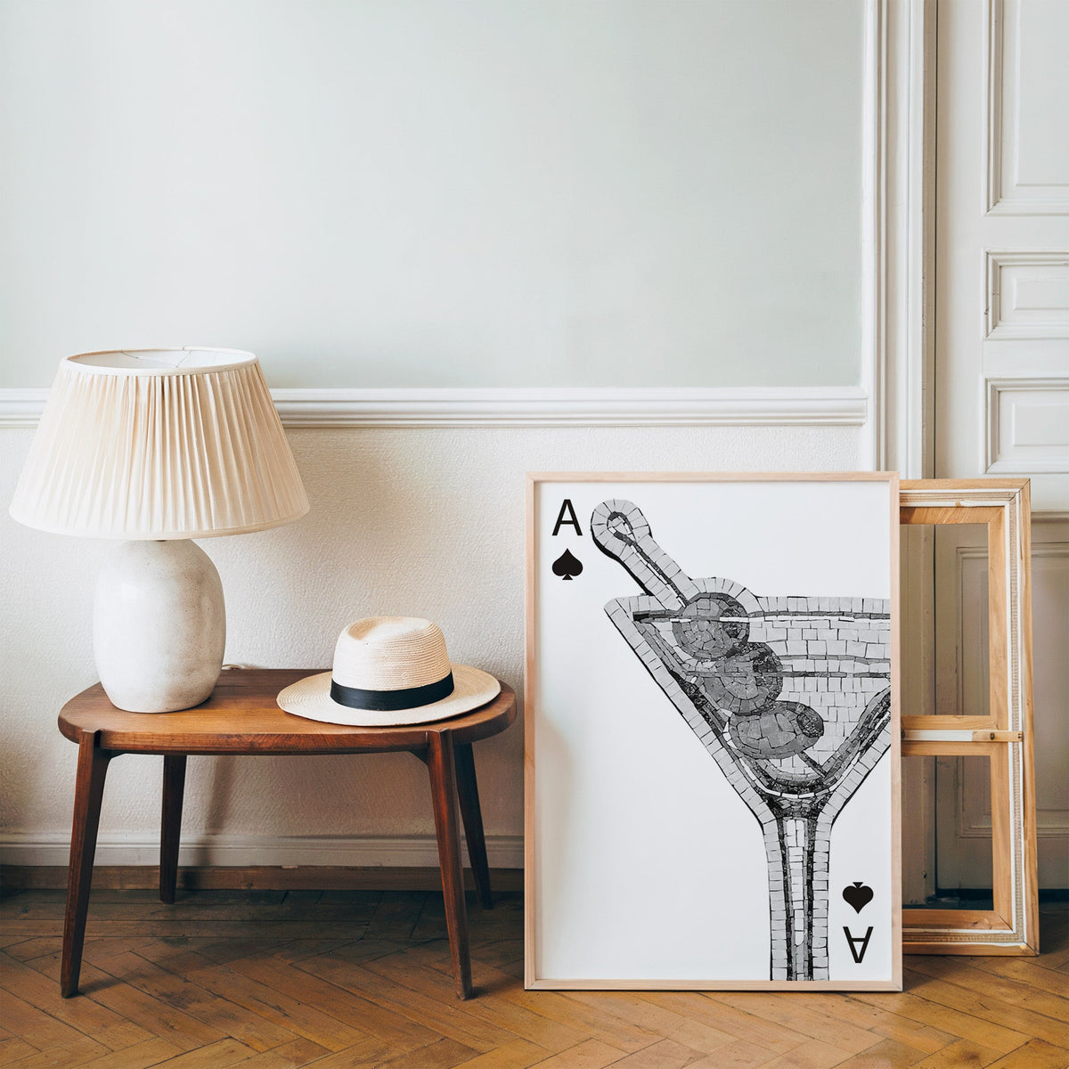 Mosaic martini glass on an ace of spades card print in a pine frame on a wood floor beside a lamp and straw hat