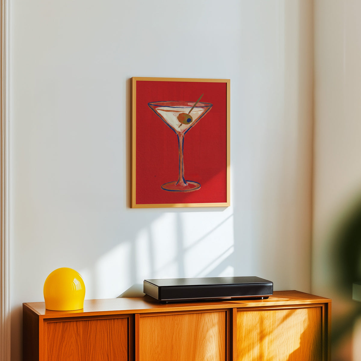 Bar cart martini print in a light oak frame above a teak sideboard with a yellow lamp