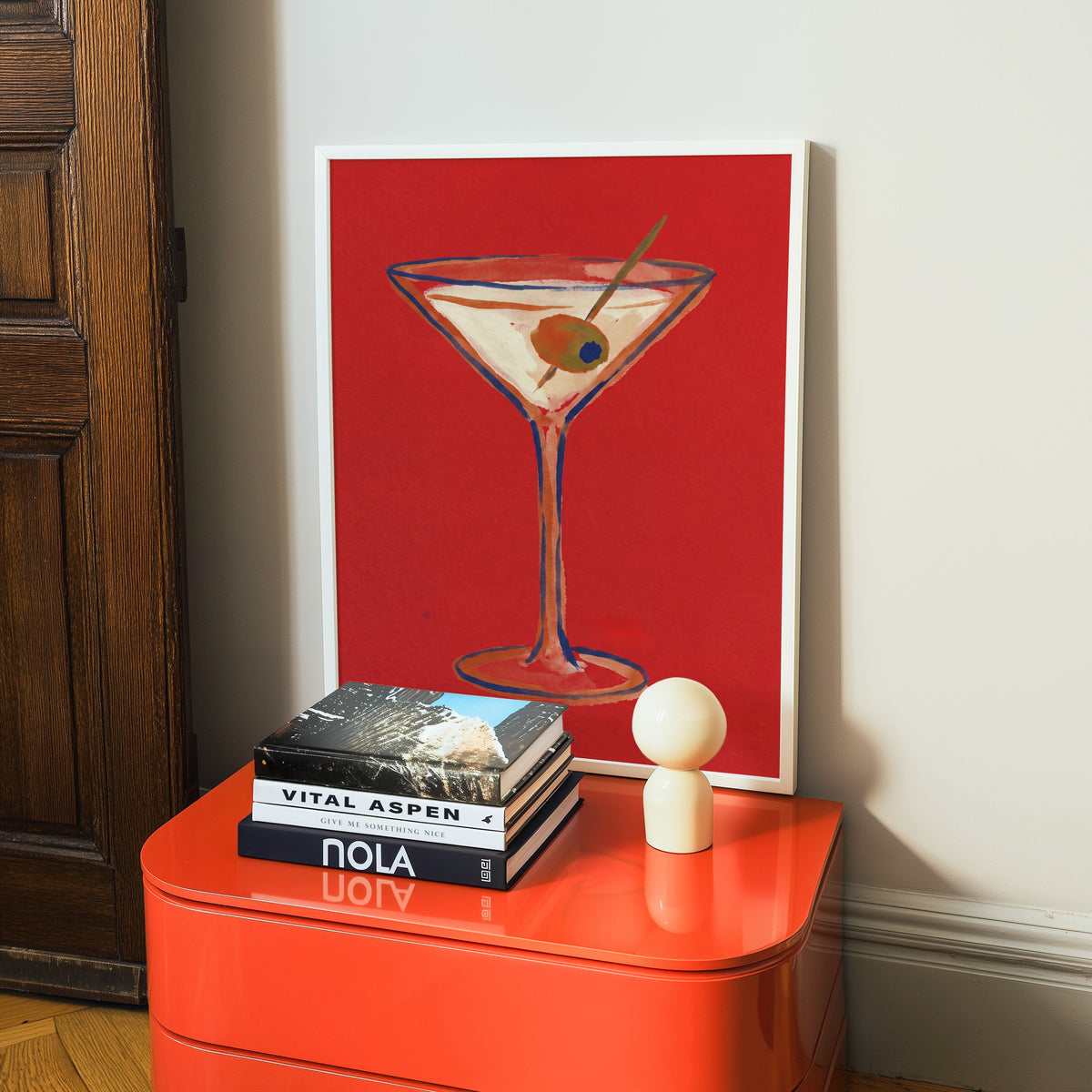 Red cocktail print in a white frame on a glossy orange cabinet with books