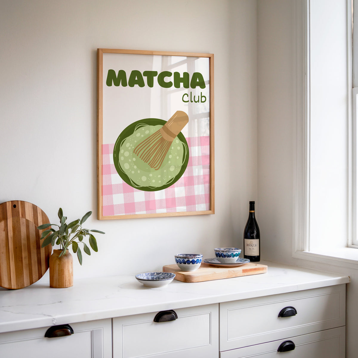 Matcha Club print in an oak frame above a white sideboard with a bottle of wine