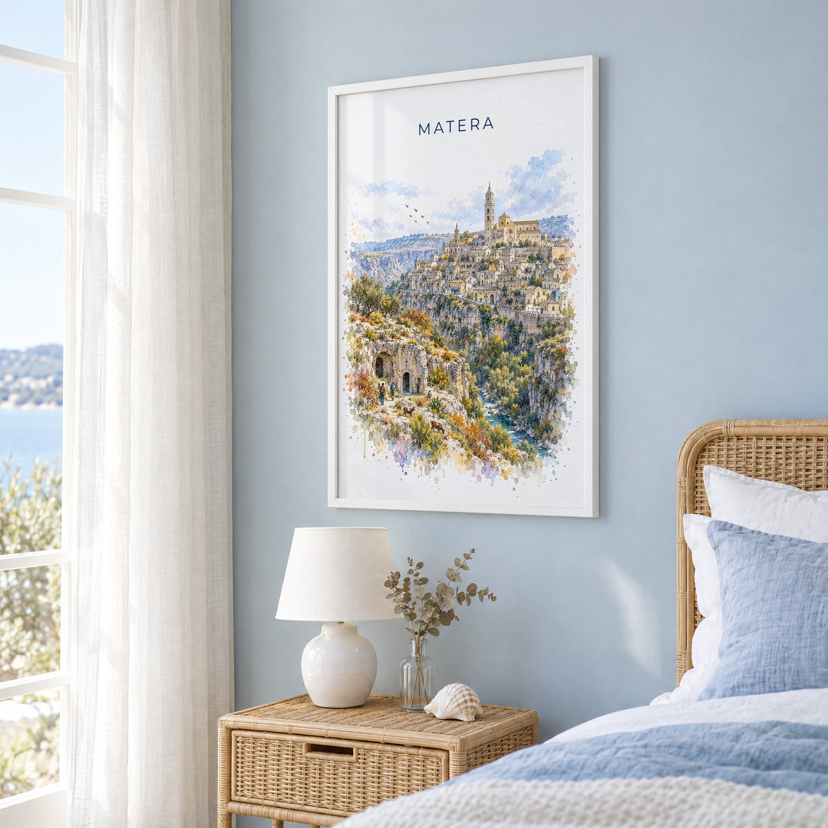 Matera watercolor print in a white frame on a pale blue wall beside a rattan bed in a coastal bedroom