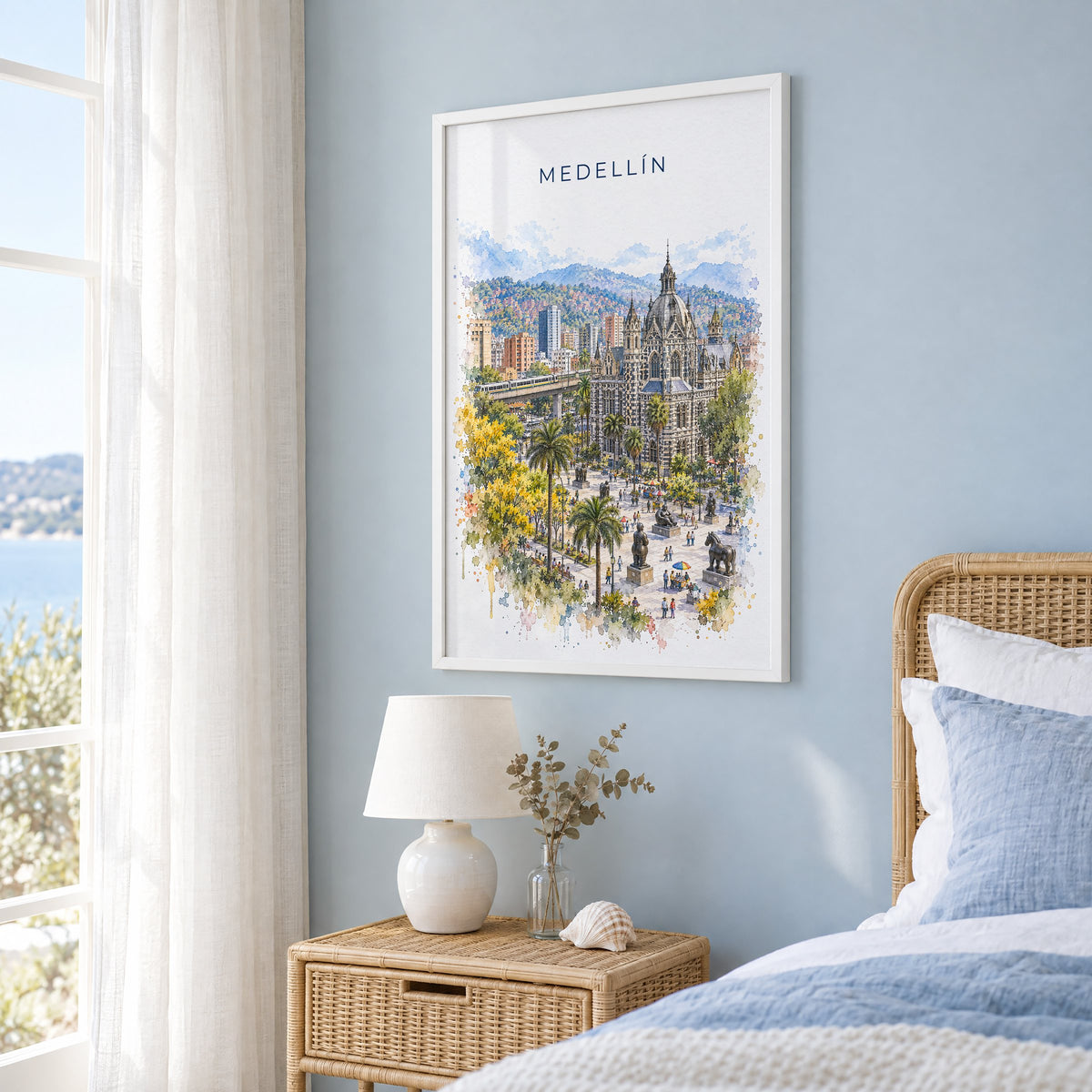 Medellín watercolor print in a white frame on a pale blue wall beside a rattan bed in a coastal bedroom