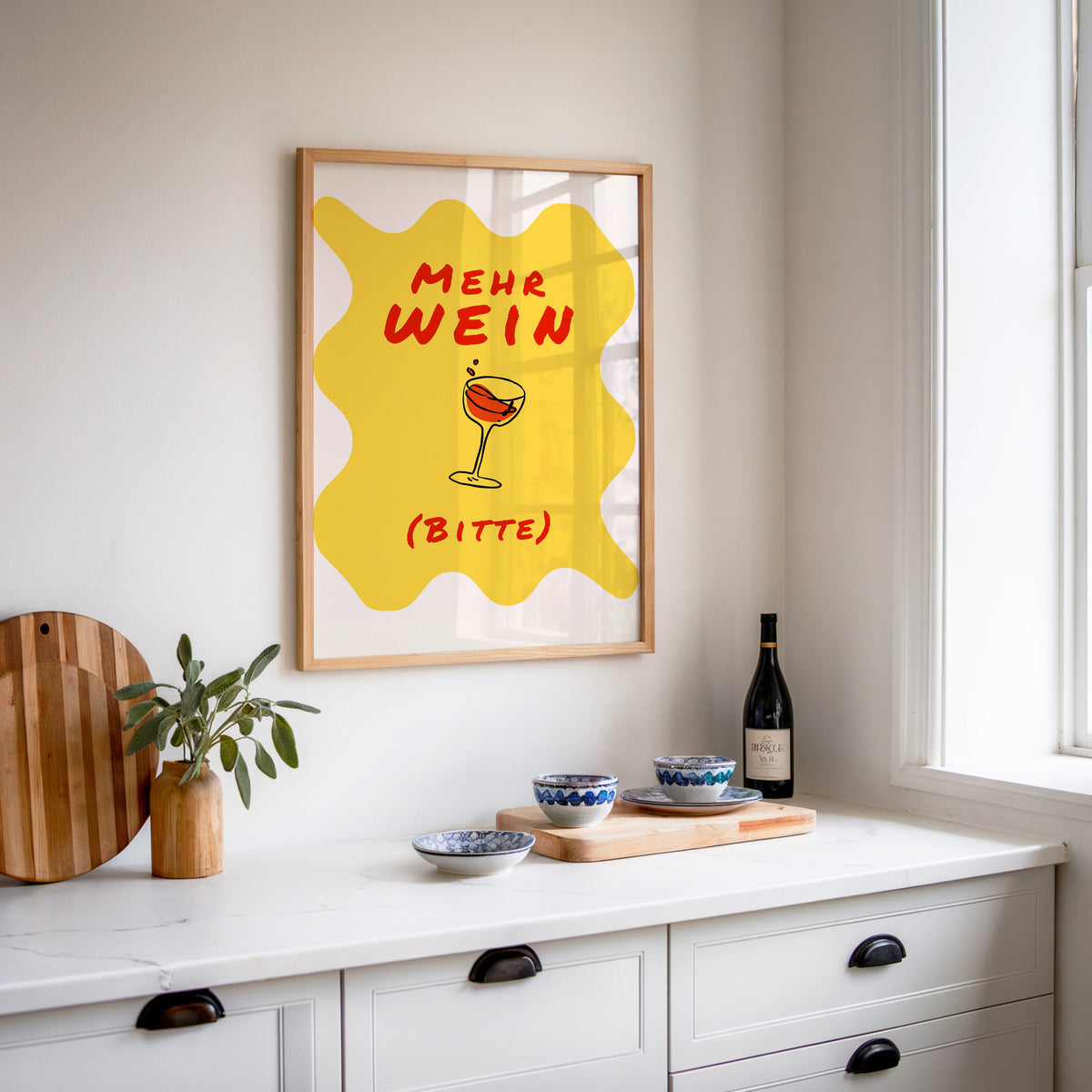 Mehr Wein Bitte print in an oak frame above a white sideboard with a bottle of wine