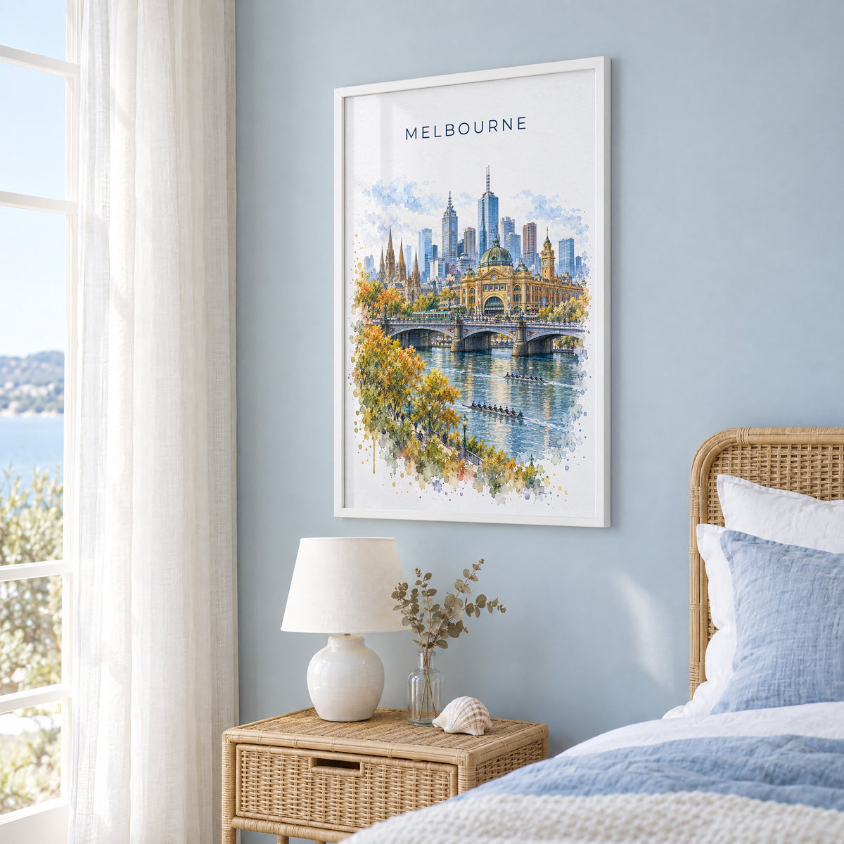 Melbourne watercolor print in a white frame on a pale blue wall beside a rattan bed in a coastal bedroom