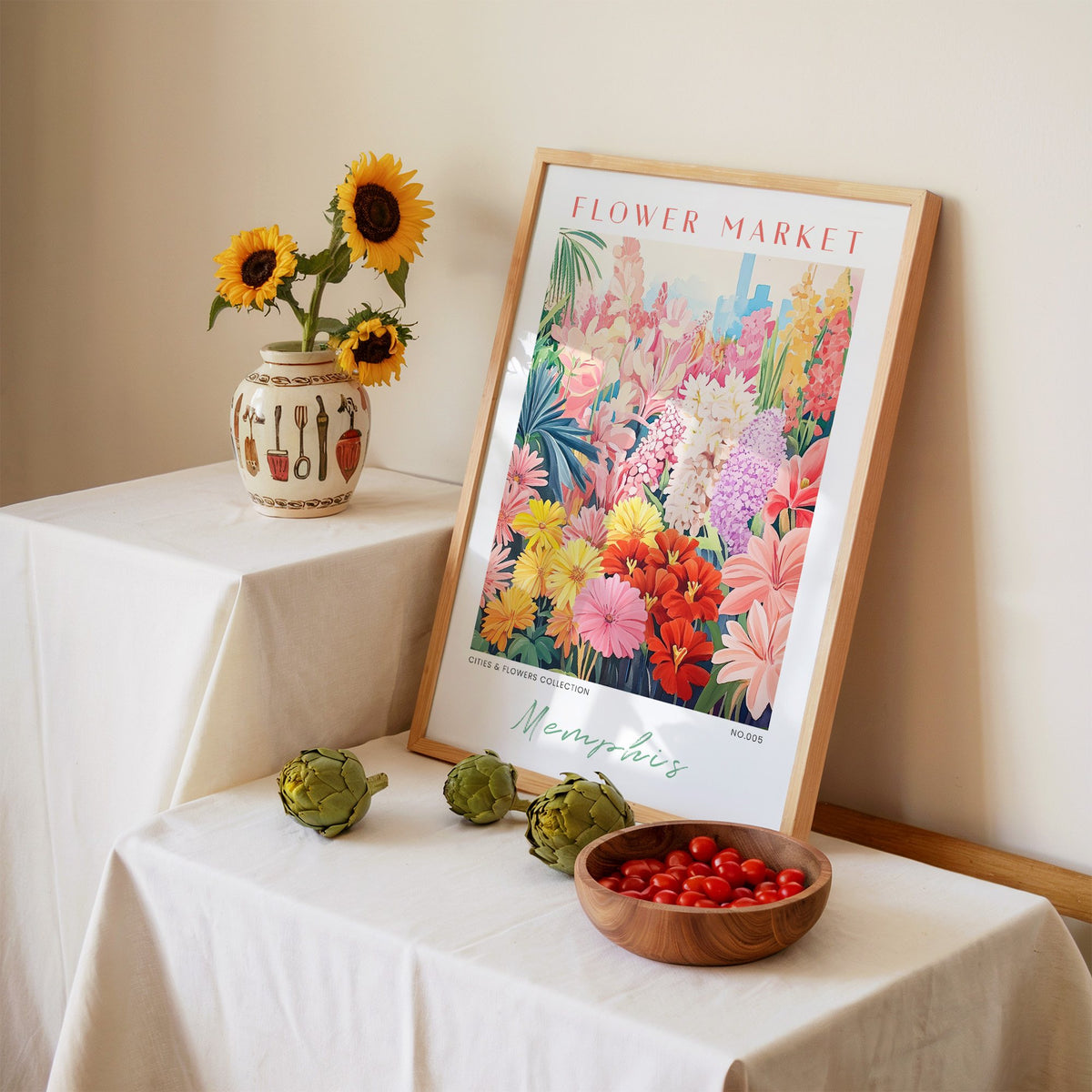 Memphis flower market print in an oak frame leaning on a linen-draped table beside a vase of sunflowers