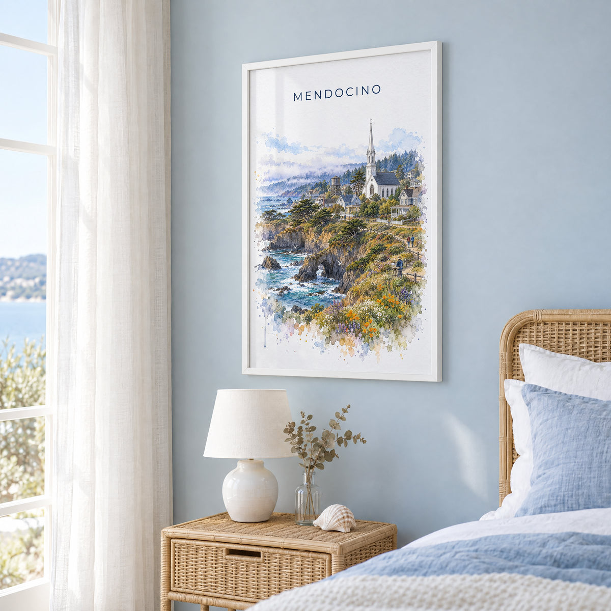 Mendocino watercolor print in a white frame on a pale blue wall beside a rattan bed in a coastal bedroom