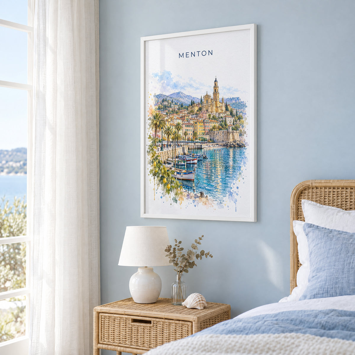 Menton watercolor print in a white frame on a pale blue wall beside a rattan bed in a coastal bedroom