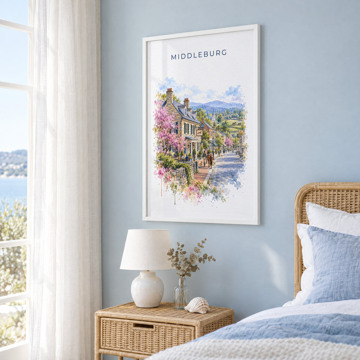 Middleburg, Virginia watercolor print in a white frame on a pale blue wall beside a rattan bed in a coastal bedroom