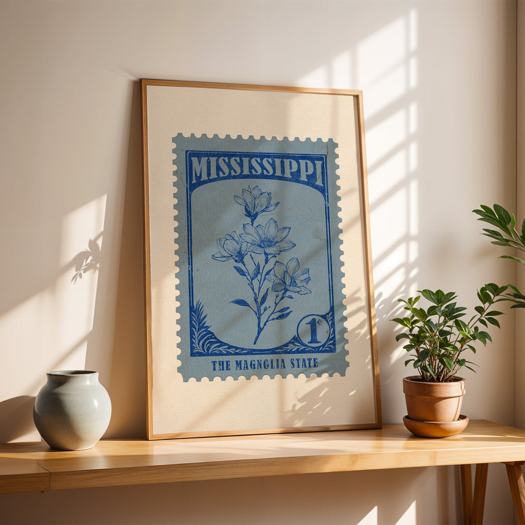 Mississippi magnolia stamp print in a light oak frame leaning on a wooden table beside a stoneware vase