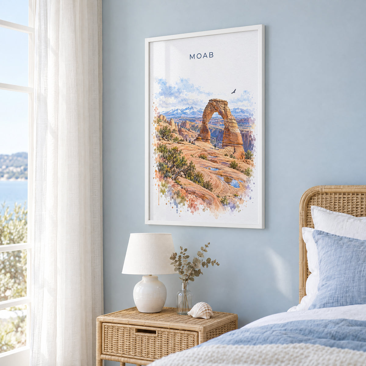 Moab watercolor print in a white frame on a pale blue wall beside a rattan bed in a coastal bedroom