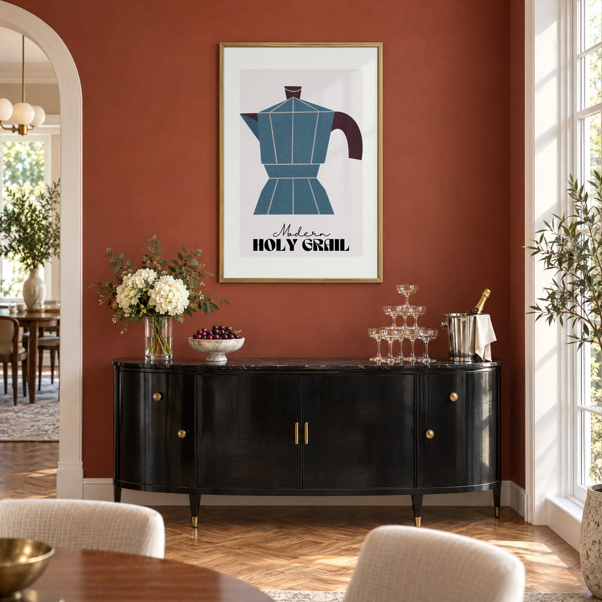 Modern Holy Grail moka pot print in a brass frame on a terracotta wall above a black buffet with a champagne tower
