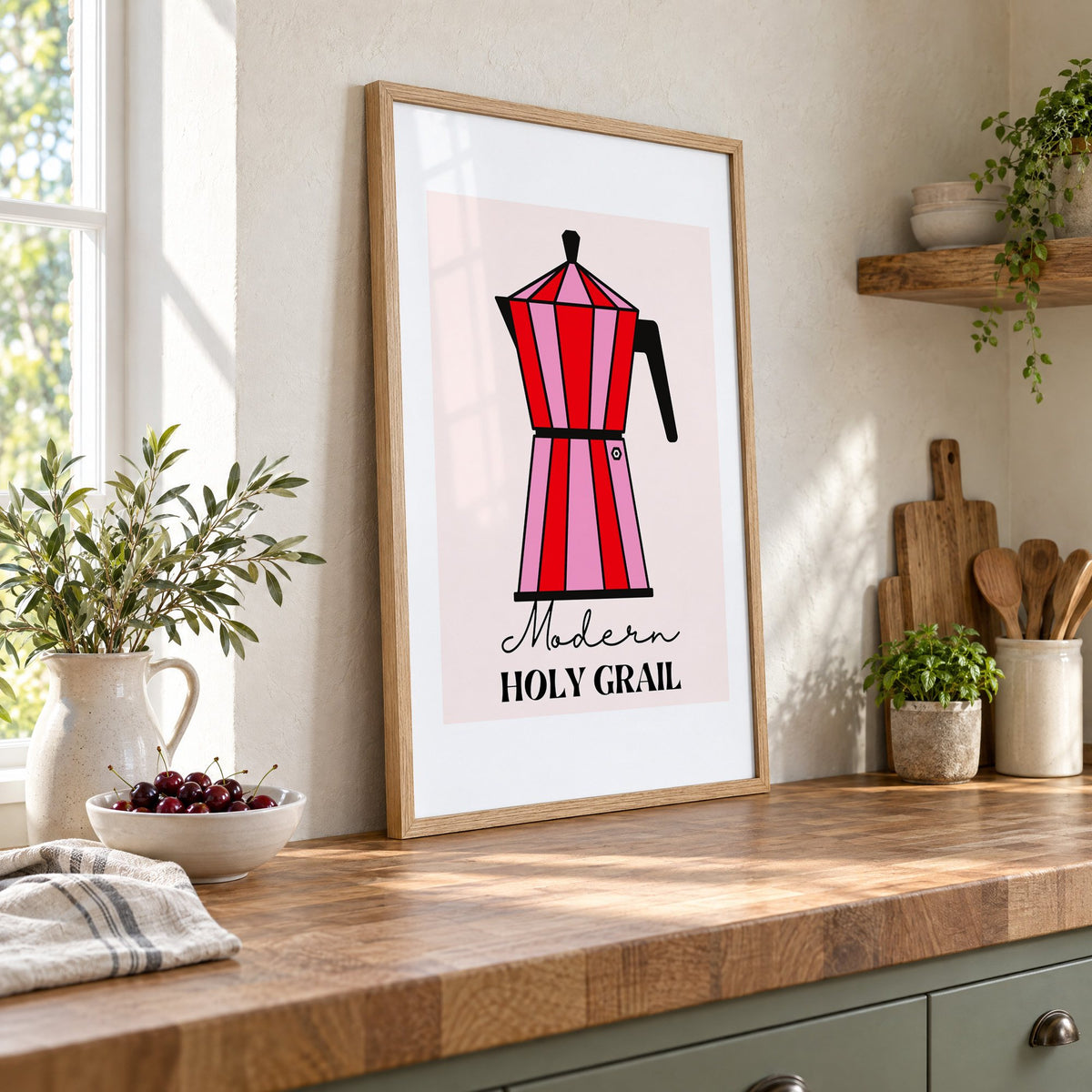 Moka pot pink and red striped print in an oak frame leaning on a butcher block island beside a bowl of cherries