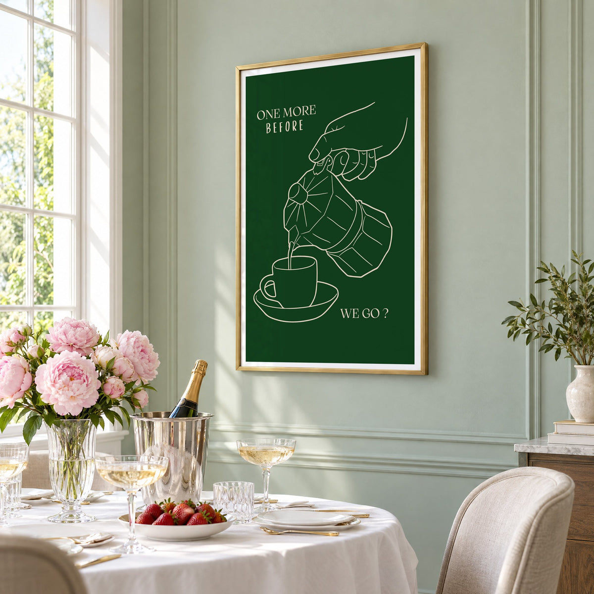 ONE MORE BEFORE WE GO? moka pot line print in a brass frame on a sage wall above a brunch table with champagne and peonies, seen at an angle