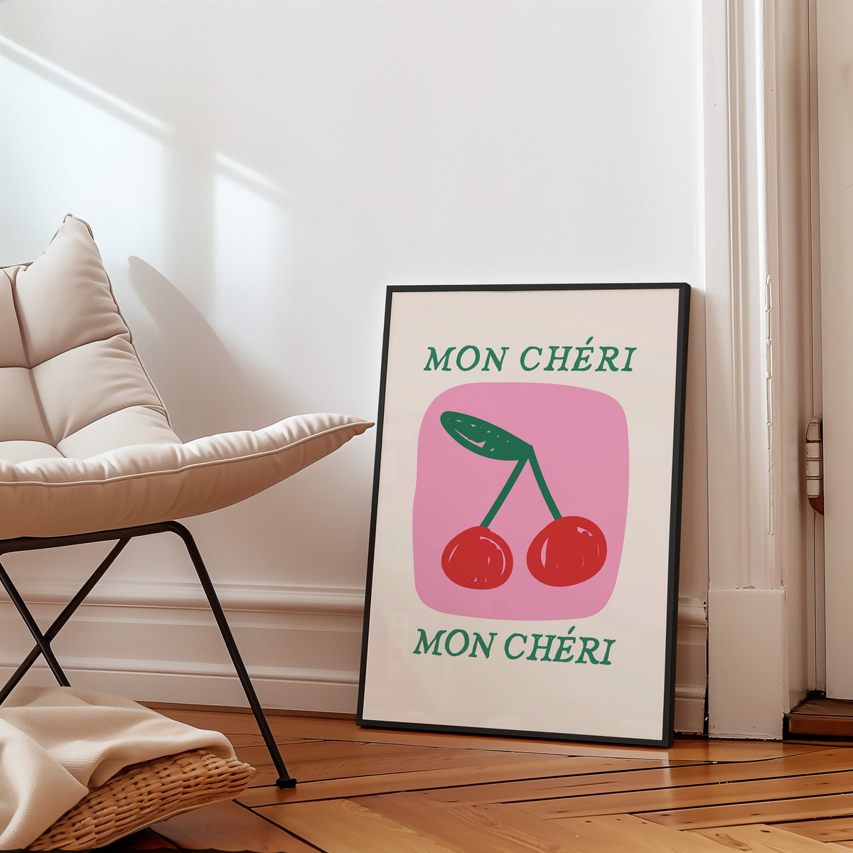 Mon Cheri two cherries pink block print in a slim black frame leaning on a herringbone floor beside a cream armchair