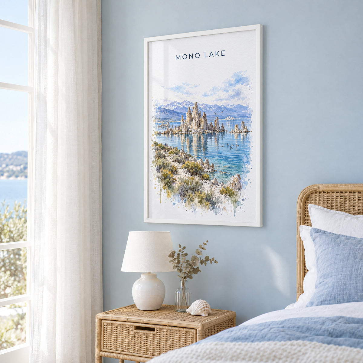 Mono Lake watercolor print in a white frame on a pale blue wall beside a rattan bed in a coastal bedroom
