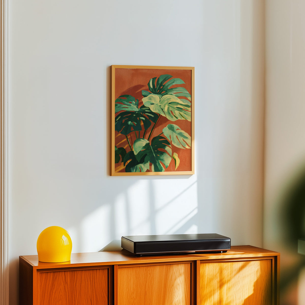 Terracotta and green leaf print in a light oak frame above a teak sideboard with a yellow lamp