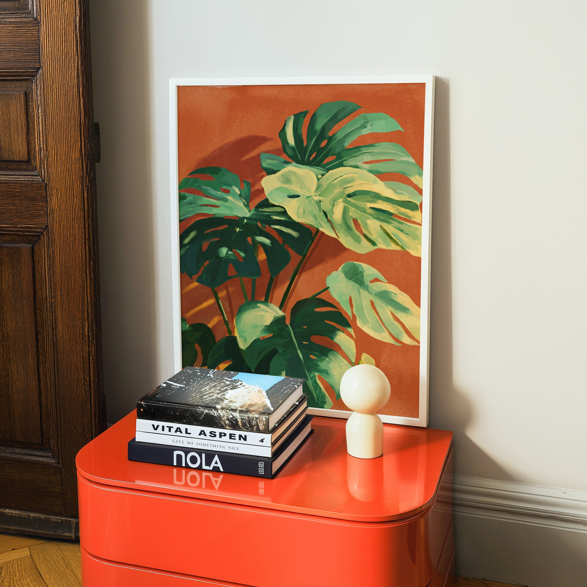 Tropical plant print in a white frame on a glossy orange cabinet with books