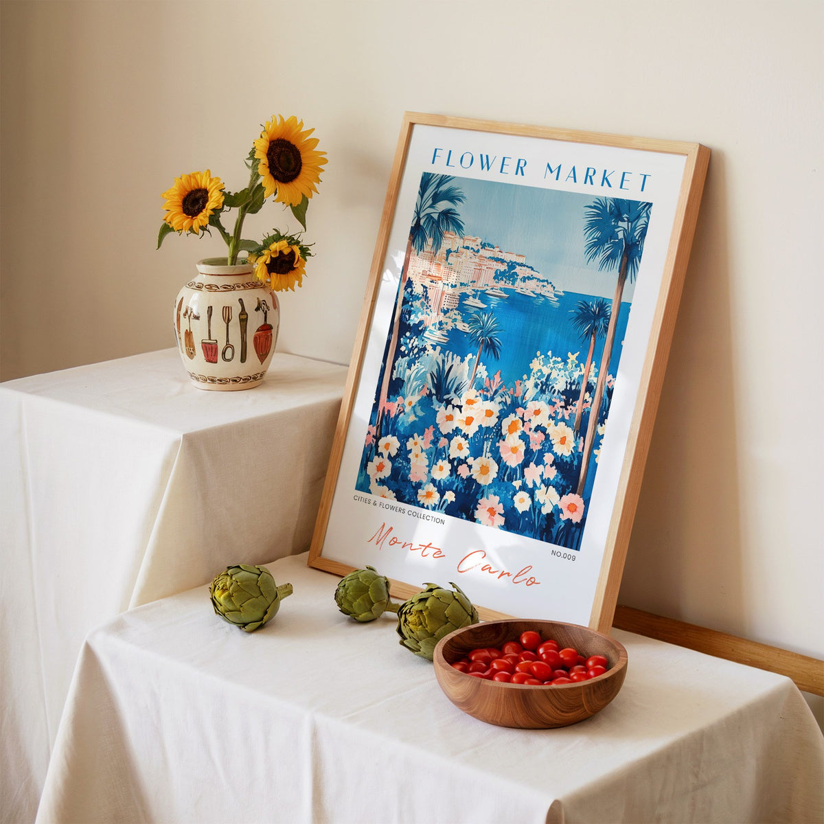 Monte Carlo flower market print in an oak frame leaning on a linen-draped table beside a vase of sunflowers