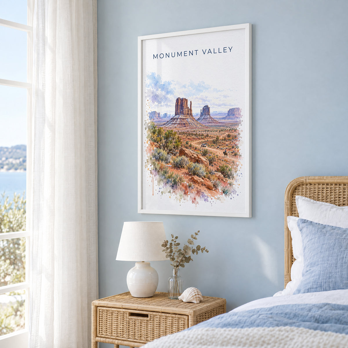 Monument Valley watercolor print in a white frame on a pale blue wall beside a rattan bed in a coastal bedroom