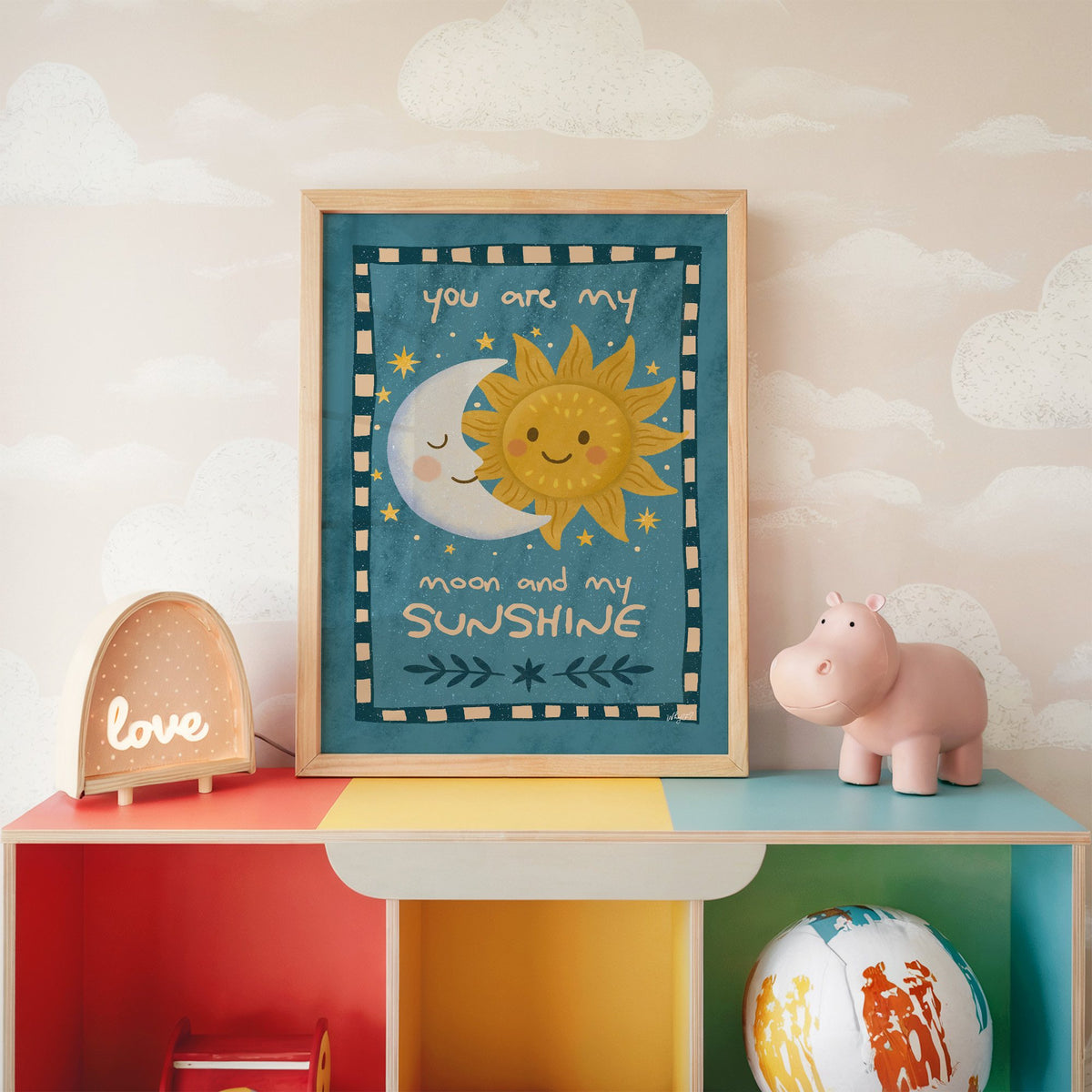 Sleepy moon and smiling sun nursery print in an oak frame on a rainbow toy shelf against cloud wallpaper