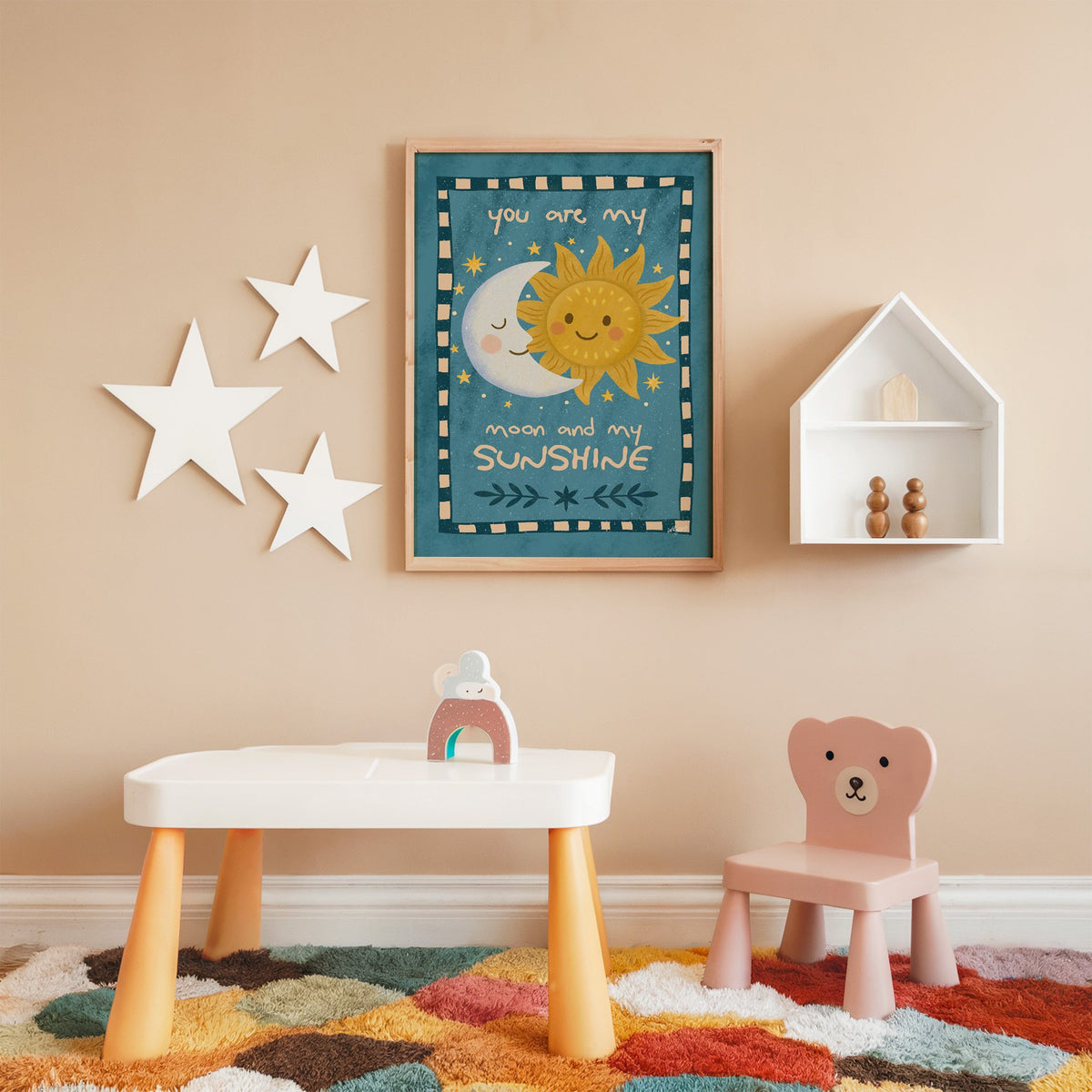 Sleepy moon and smiling sun nursery print in an oak frame on a peach wall above a kids table with wall stars