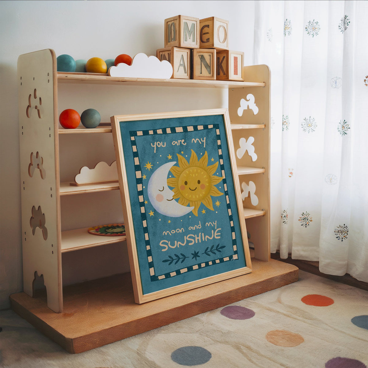 Sleepy moon and smiling sun nursery print in an oak frame leaning on a toy shelf with wooden letter blocks