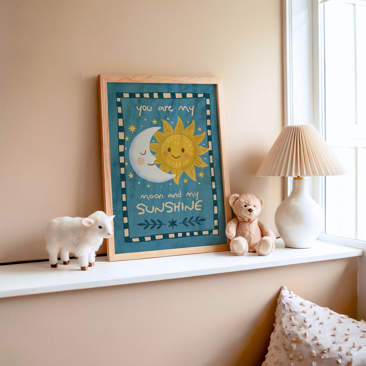 Sleepy moon and smiling sun nursery print in an oak frame on a window ledge with a toy lamb and teddy