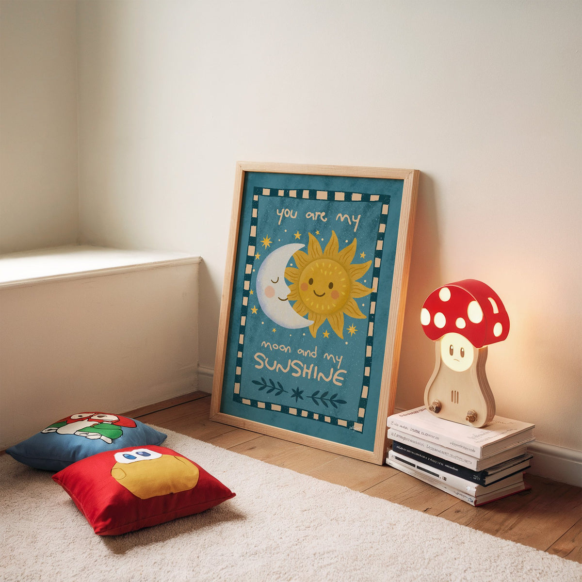 Sleepy moon and smiling sun nursery print in an oak frame leaning on a rug beside a mushroom lamp and floor cushions