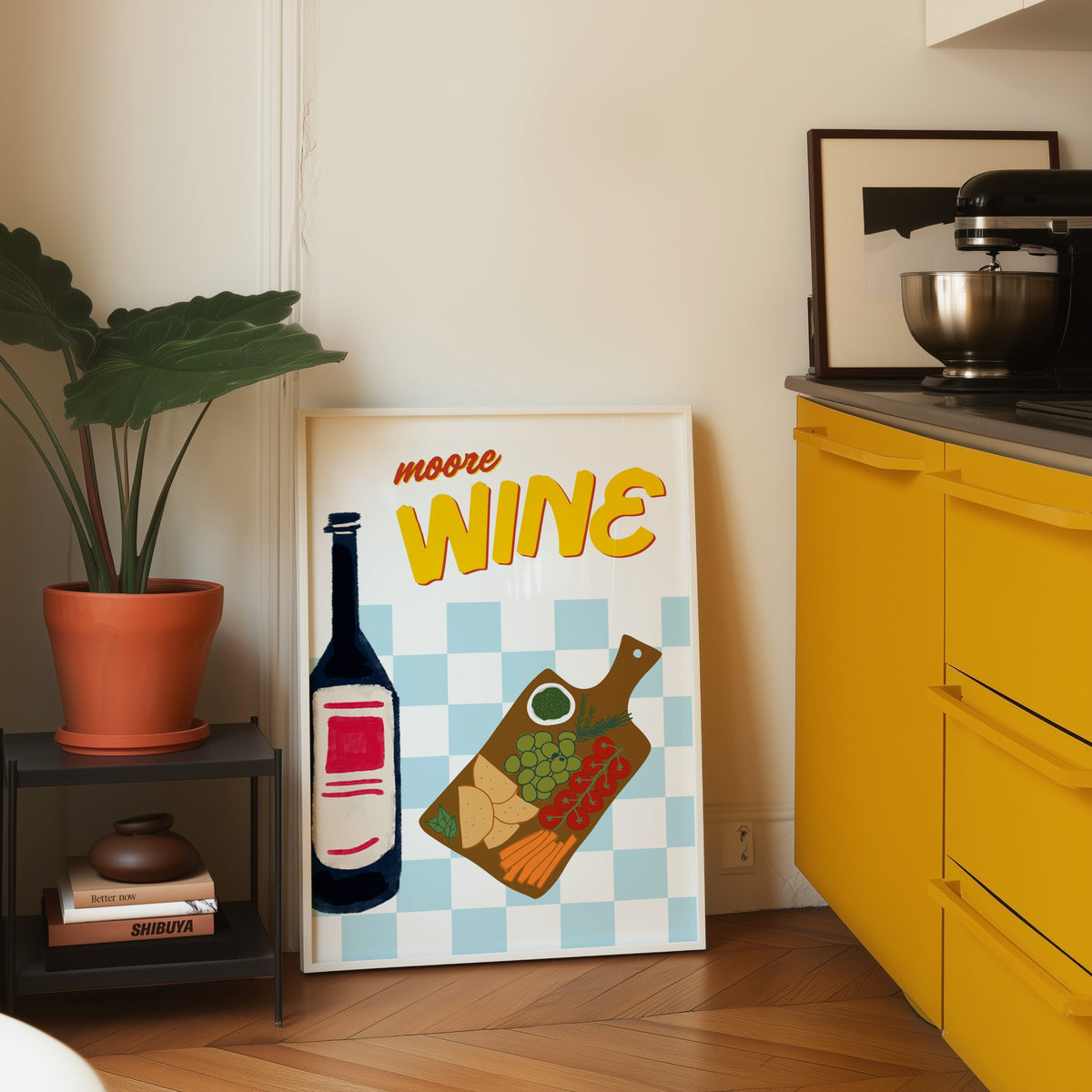 Wine and cheese board print in a white frame leaning on parquet beside a bright yellow kitchen