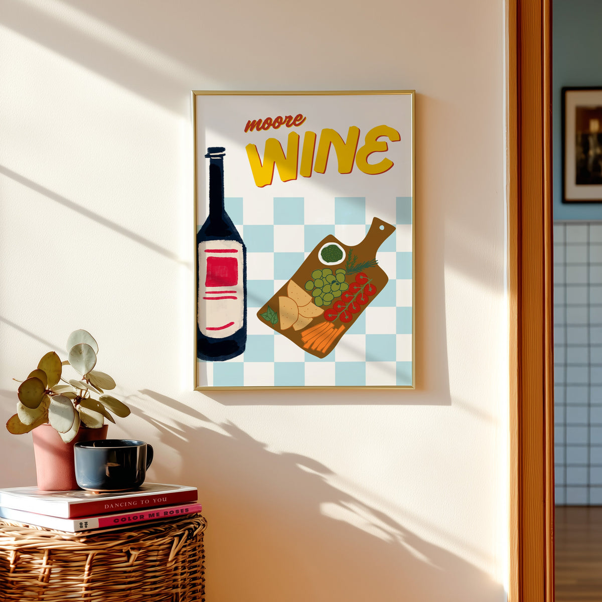 Wine and cheese board print shown in a gold frame above a basket of books and a pilea plant