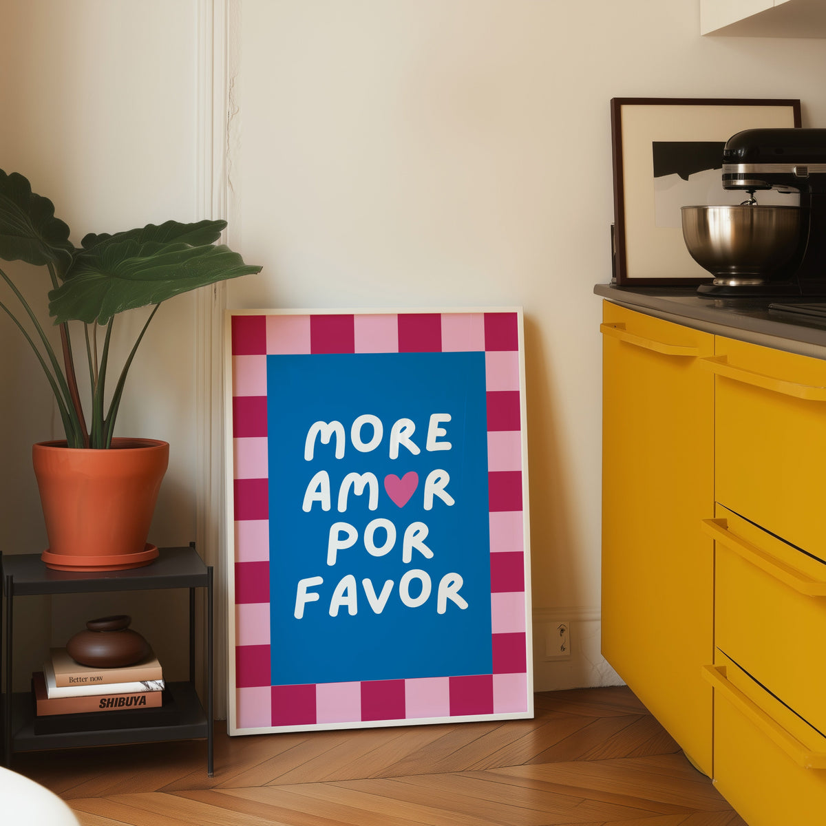 More Amor Por Favor print in a white frame leaning on parquet beside a bright yellow kitchen