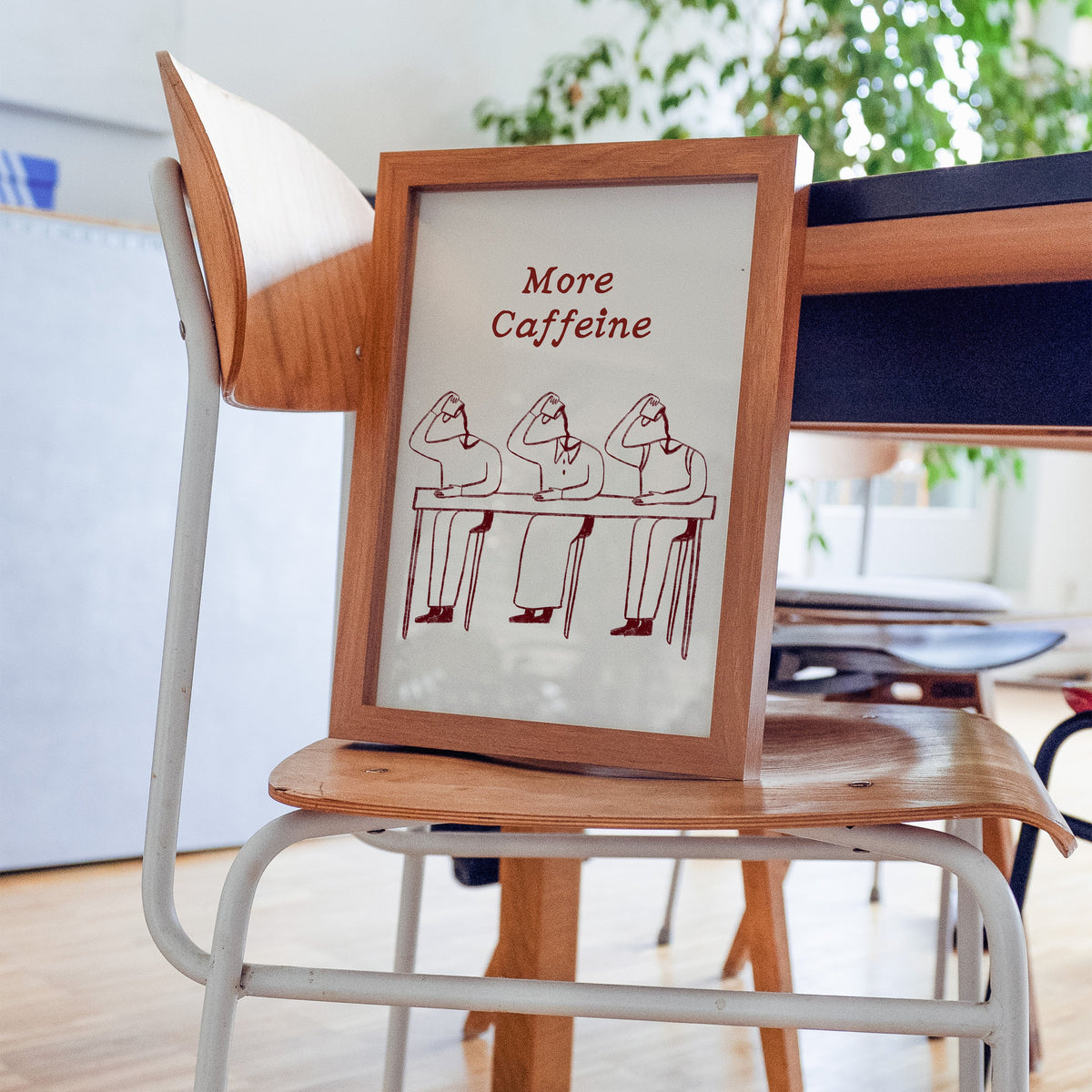 More Caffeine desk slump print in a walnut frame resting on a plywood studio chair beside a dark desk