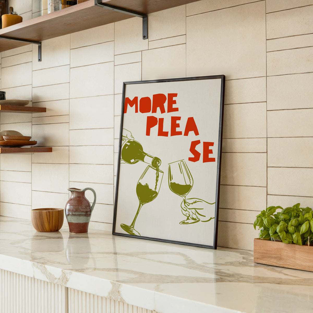 More Please wine pour print in a black frame leaning on a marble counter in a tiled kitchen