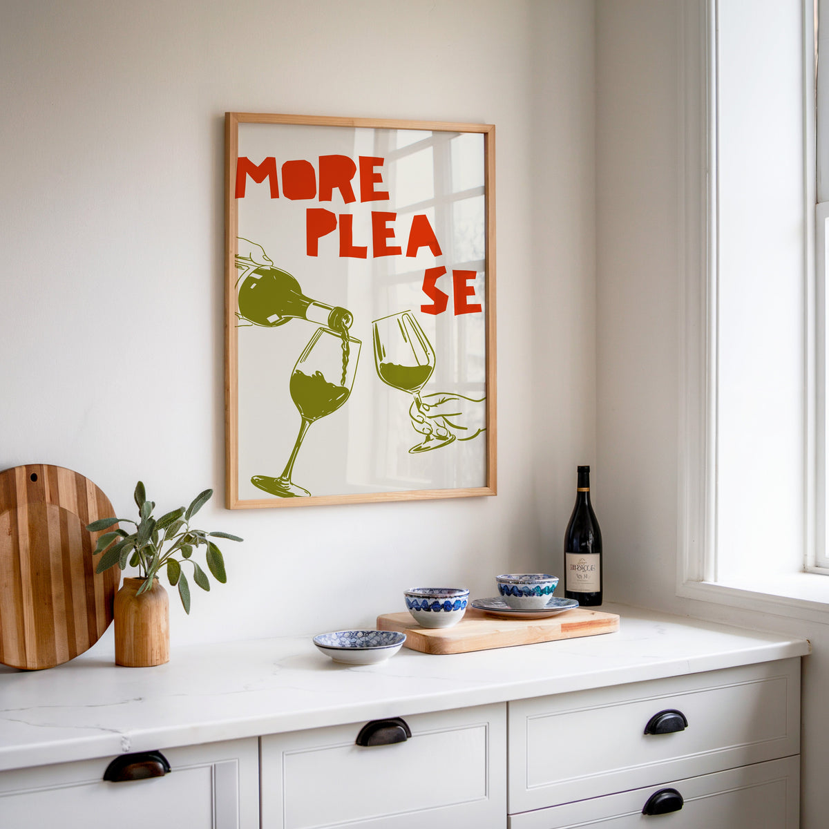 More Please wine pour print in an oak frame above a white sideboard with a bottle of wine