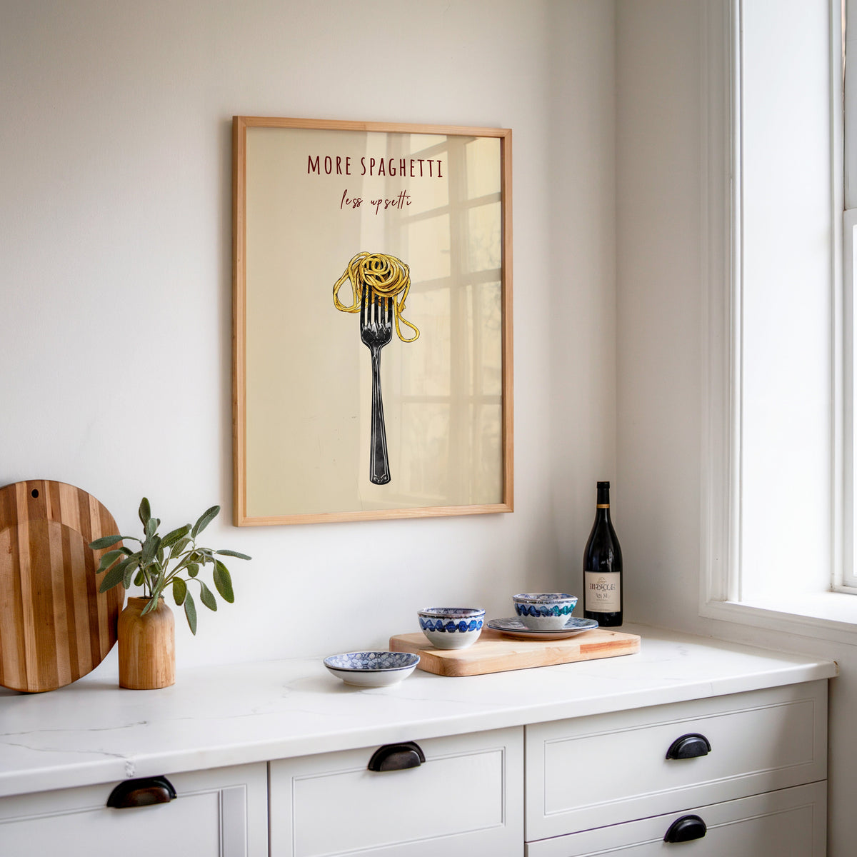 More Spaghetti Less Upsetti print in an oak frame above a white sideboard with a bottle of wine