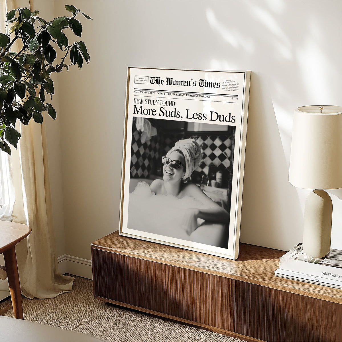 Woman in bubble bath mock newspaper print in a white frame on a walnut sideboard beside a ceramic lamp