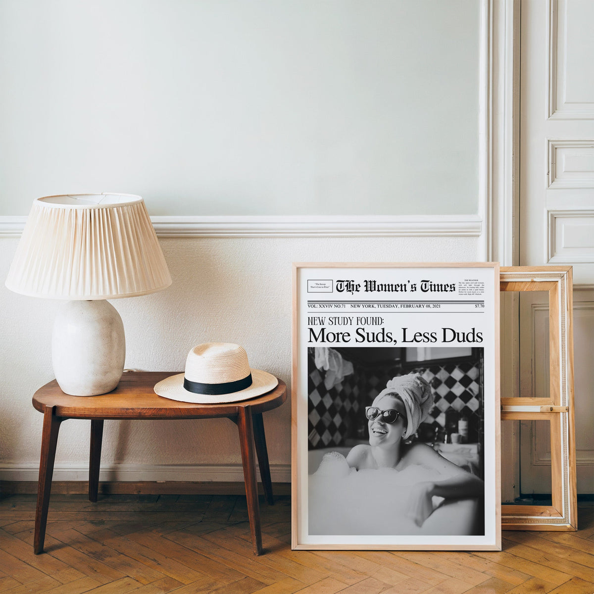 Woman in bubble bath mock newspaper print in a pine frame on a wood floor beside a lamp and straw hat