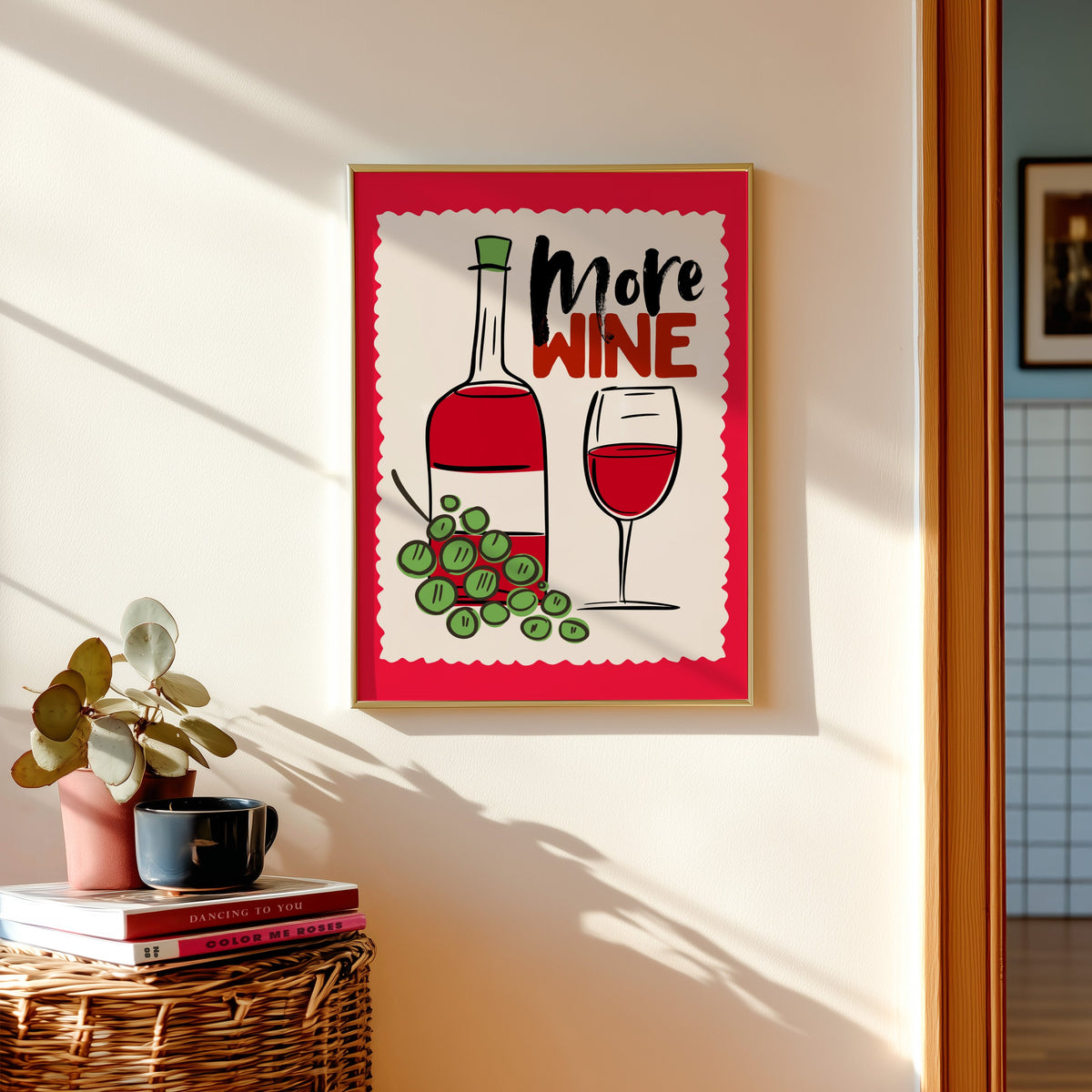 More Wine print in a gold frame above a basket of books and a pilea plant