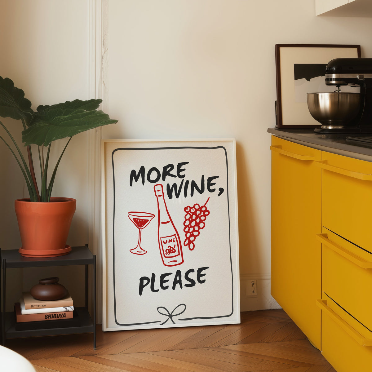 More Wine Please print in a white frame leaning on parquet beside a bright yellow kitchen