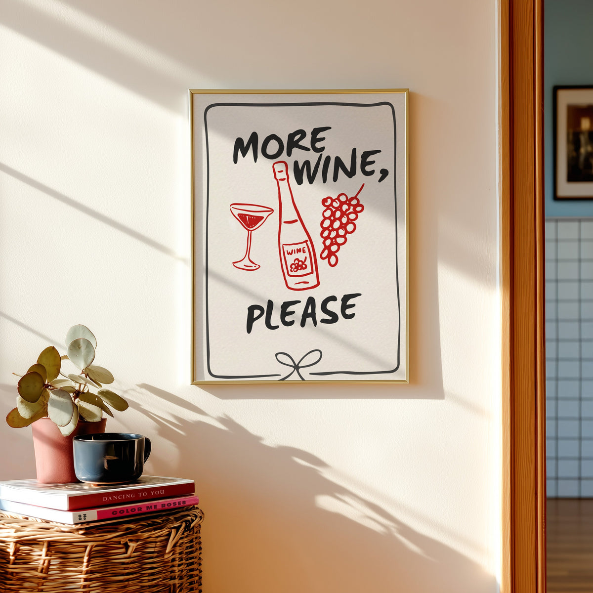 More Wine Please print shown in a gold frame above a basket of books and a pilea plant