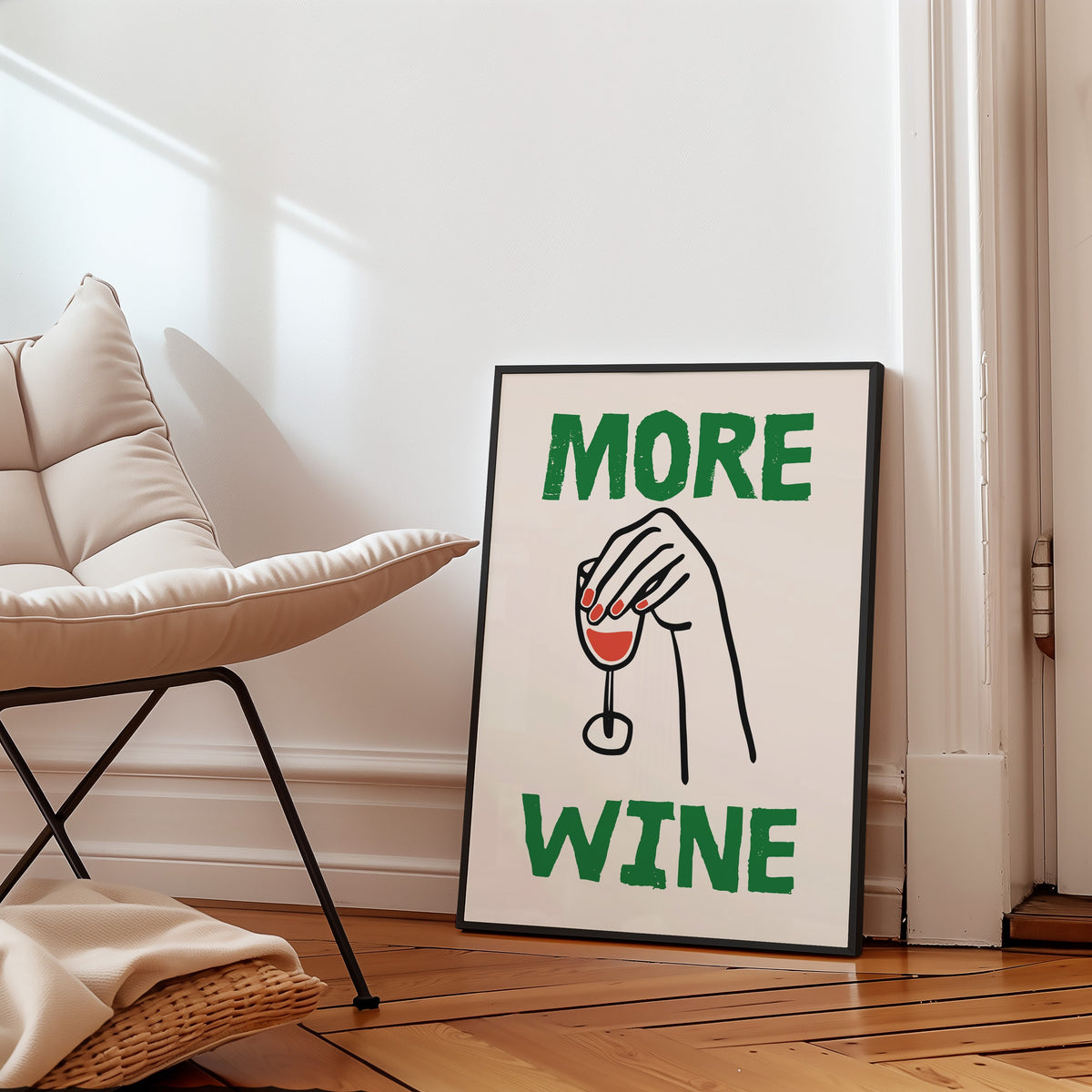 More Wine upside down glass print in a slim black frame leaning on a herringbone floor beside a cream armchair