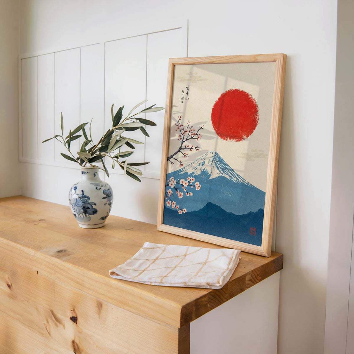 blue Mount Fuji with red sun and sakura print in a pine frame on a wooden counter beside a blue and white porcelain vase
